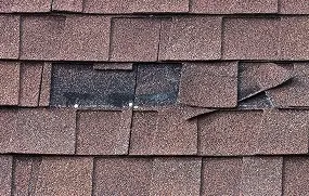 Close-up of brown roof shingles with a broken and missing section revealing the underlying material.