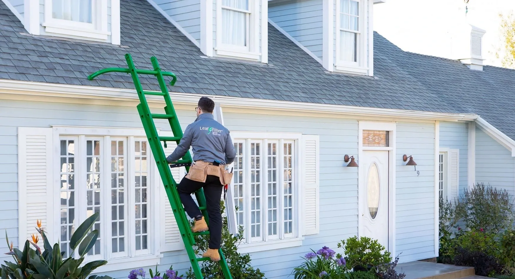 A technician in a LeafFilter uniform climbs a green ladder to work on a light blue house with white trim.